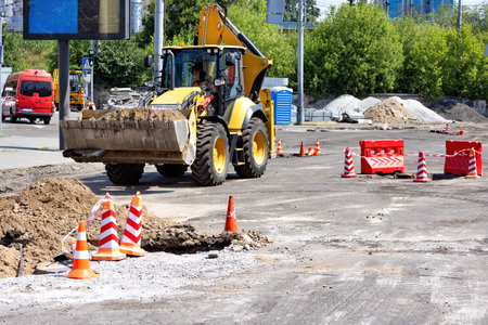 Heavy machinery is actively involved in road repairs under a clear blue sky. Traffic cones are...