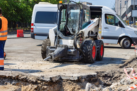 A construction vehicle diligently works on a city street, removing concrete while workers oversee the activity.の写真素材
