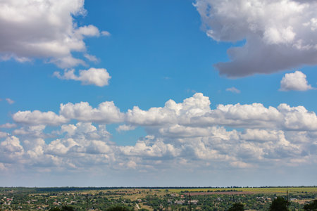 Fluffy white clouds drift across a vivid blue sky, casting playful shadows on a sprawling green landscape below. The scene captures a peaceful moment of nature's beauty during midday. Copy space.の写真素材