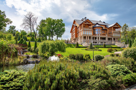 A grand mansion stands majestically amidst manicured gardens, featuring elegant landscaping, a tranquil pond, and a serene waterfall, all under a bright, blue sky.の写真素材