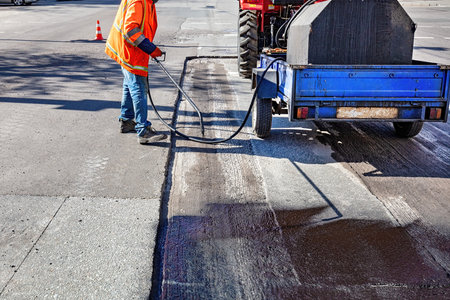 A worker in an orange safety vest operates a sealant machine to coat a recently paved section of road. This activity takes place mid-morning, highlighting the busy urban environment.の写真素材