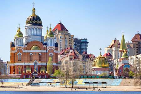 Under a clear blue sky, the colorful domes and spires of an Orthodox church tower majestically above the riverbank. People stroll along the embankment, admiring the stunning buildings and enjoying a sunny day in the city.の写真素材