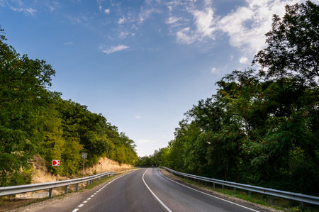 Road turn. Landscape highway, forest, mountains and sky on the horizon. Empty long mountain road to the horizon on a sunny summer day with dramatic cloudy sky. Highway turn panoramic view.の写真素材