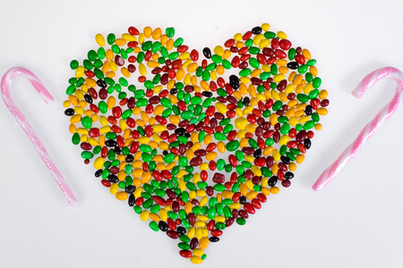 Coloured jelly beans are scattered in the shape of heart and sweet pink and white striped cane on a white background.の写真素材