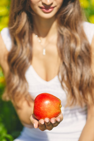 Young lady with long hair holding a red apple and offer it to you on the field of dandelionsの写真素材