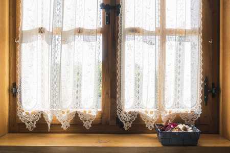 a wooden window with a white embroidered curtain and a small basket of dried flowers on the windowsillの写真素材