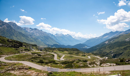 Winding alpine road with sharp curves leading through the mountains in Switzerland, surrounded by green valleys and distant peaks under a bright summer skyの写真素材