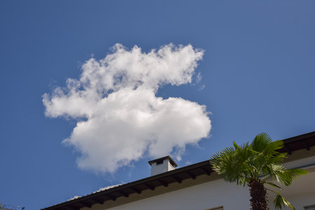 A fluffy white cloud appears to rise like smoke from a chimney against a vivid blue sky, creating a playful optical illusionの写真素材