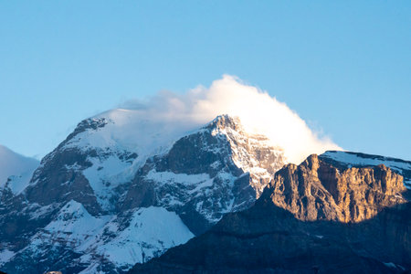 Snow-covered peak of Mount TÃ¶di in Switzerland, illuminated by golden sunlight, with dramatic clouds drifting over the Alpine summitの写真素材