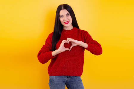 Young woman in a warm sweater shows hands a heart on a yellow background. Christmas celebration.の写真素材