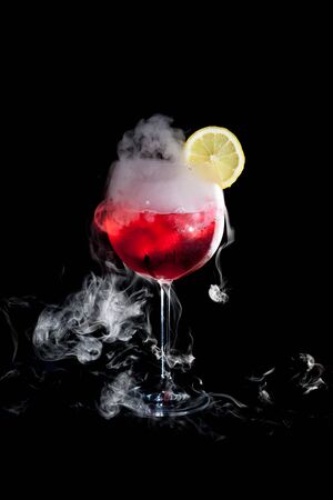 A cup of red cold cocktail with dry ice smoke and a lemon slice and black backgroundの写真素材