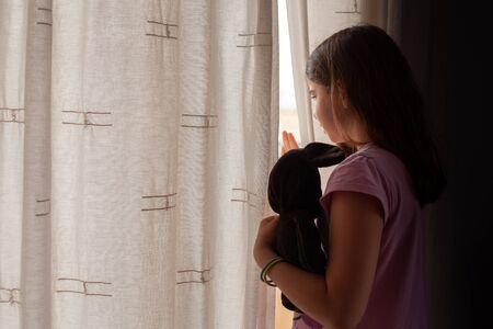 Lockdown concept. Little girl with her teddy bear looking at the street between the window curtains. Stay at homeの写真素材