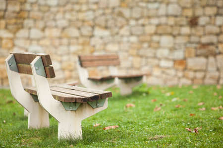 Empty street benches without people in a city park on a blurred background of an old wall made of natural stone. Autumn fallen leaves on a green lawn. Soft daylight, cloudy weather.の写真素材