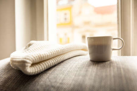 Image of a coffee cup on a soft pillow on a windowsill with a white knitted hat overlooking the...
