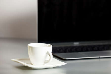 Opened laptop and coffee cup on saucer in the foreground on the gray table and white background. Remote workplace, home office. Online communication. Moke up with laptop and coffe cup.の写真素材