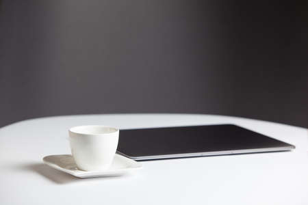Composition with a modern closed laptop and coffee cup with saucer on the white table on a gray...