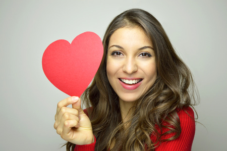 Valentine's Day. Beautiful young woman wearing red dress and holding a red heart paper on gray background. Valentine concept.の写真素材