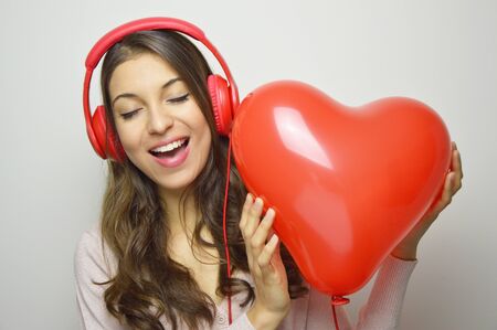 Valentine's day love girl concept. Cute enthusiast girl with red headphone listening to the music of his heart. Young happy woman holding and listening to her red balloon heart.の写真素材