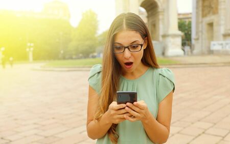 Pretty astonished business woman looks her smart phone in the square.