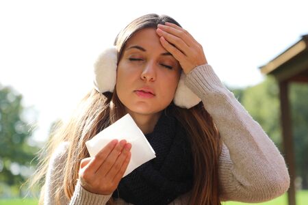 Young woman with earmuffs and scarf suffering migraine headache holding tissue outdoors.の写真素材