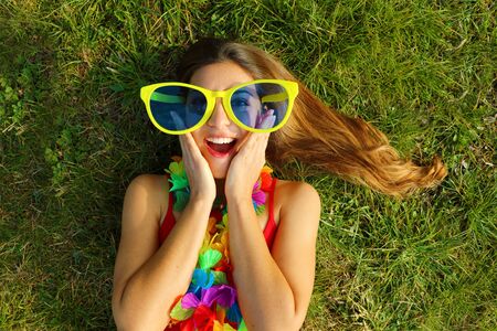 Surprised girl wake up on grass after Carnival party. Happy excited oung woman with carnival garland and big funny sunglasses lying on grass.の写真素材
