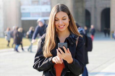 Happy beautiful young high school girl with smart phone outdoors on sunny spring day texting and smilingの写真素材