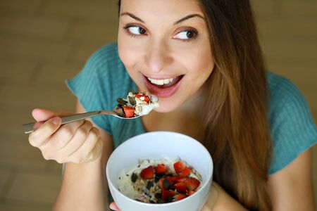 Close up view from above of beautiful young woman eating skyr yogurt with strawberries, cereal muesli and seeds at home, looking to the side, focus on model eyes, indoor picture. Healthy food concept.の写真素材