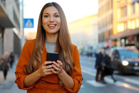 Happy beautiful young woman looking at camera and holding mobile phone outdoor with blurred people on street background. City lifestyle people technology.の写真素材
