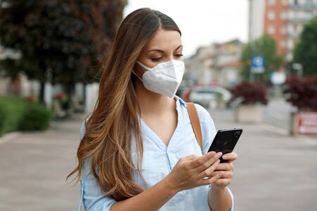 COVID-19 Young Woman Wearing KN95 FFP2 Mask Using Smart Phone Application Software in City Street to Aid Contact Tracing and Self Diagnostic in Response to the Pandemic Coronavirus 2019の写真素材