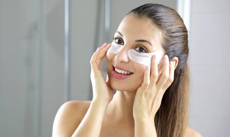 Smiling beautiful woman applying anti-fatigue under-eye mask looking herself in the mirror in the bathroom. Skin care girl touch patches of fabric mask under eyes to reduce eye bags.の写真素材