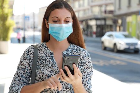 Portrait of young beautiful business woman wearing surgical mask typing on mobile phone in city streetの写真素材