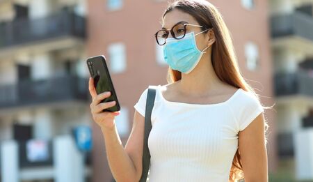 Beautiful young woman with medical mask messaging on the smart phone in city street. Pretty girl having smart phone conversation outdoor.の写真素材