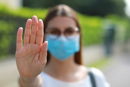 STOP COVID-19 Unidentified Woman Wearing Surgical Mask Showing STOP Gesture Looking at Camera Outdoors. Focus on open hand palm.の写真素材