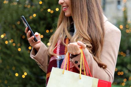 Portrait of smiling young shopper woman buying online on smartphone in city street with christmas...