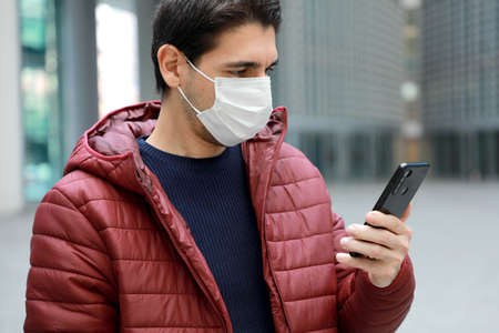 COVID-19 Portrait of young man wearing protective mask reading news on smartphone app in modern city...