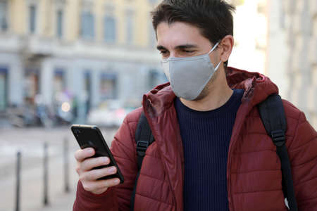 Happy young man with protective mask looks his smartphone in street. Student guy with face mask using mobile phone outdoors.の写真素材