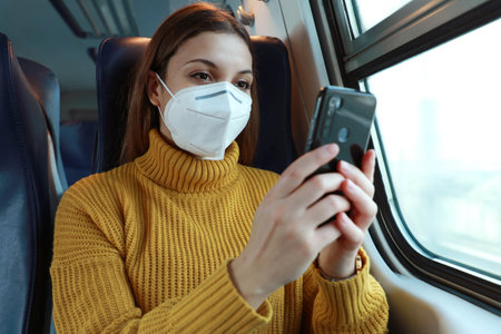 Relaxed woman with KN95 FFP2 face mask using smart phone app. Train passenger with protective mask traveling sitting in business class texting on mobile phone. Travel safely on public transport.の写真素材
