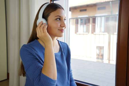 Beautiful young woman at home enjoys listening music with wireless headphones looking through the windowの写真素材