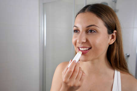 Smiling young woman applying lip balm in front of the mirror indoorの写真素材