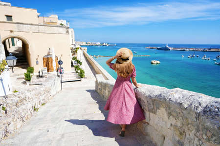 Beautiful young woman with hat walking along the ancient walls of Otranto looking at stunning panoramic view of Otranto village in Salento, Italyの写真素材