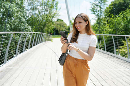 Happy young woman with comfortable clothes typing with finger on smart phone on city park. Sustainable eco-friendly city and lifestyle technology concept.の写真素材