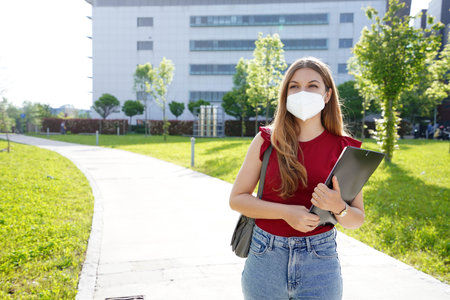 Business girl with protective mask walks holding folders in her hands with office buildings on the backgroundの写真素材