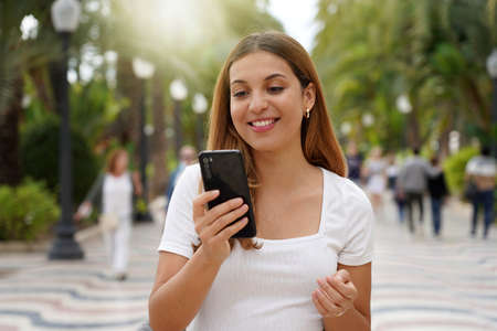 Excited young woman watching her smartphone when walking in the street with blurred people on the...