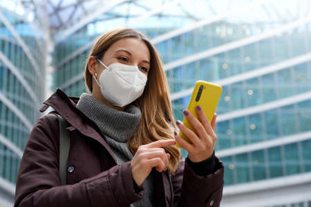 Portrait of woman wearing protective medical face mask using a phone with modern city background. Low angle.の写真素材