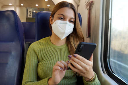 Relaxed woman on train wearing medical face mask using smart phone app. Commuter with protective...