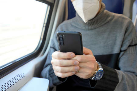 Man on public transport using mobile app wearing medical face mask. Train commuter holding cellphone...