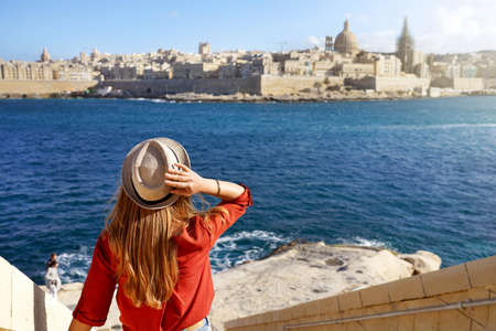 Beautiful young woman with hat descends stairs in Malta towards waterfront looking at panoramic view...