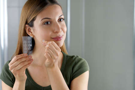 Beautiful young woman applying acne treatment anti-pickel patch on a pimple in bathroom at homeの写真素材