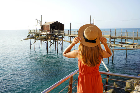 Romantic sunset in Italy. Back view of young woman with hat and dress enjoying view of Trabucco an...