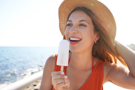 Close-up of beautiful girl with hat holding a white ice pop on the beach looking to the sideの写真素材